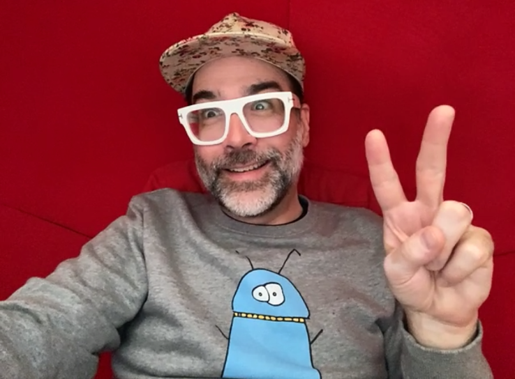 Caucasian man (Bleu) with grey beard in casual clothing holding up a peace sign.