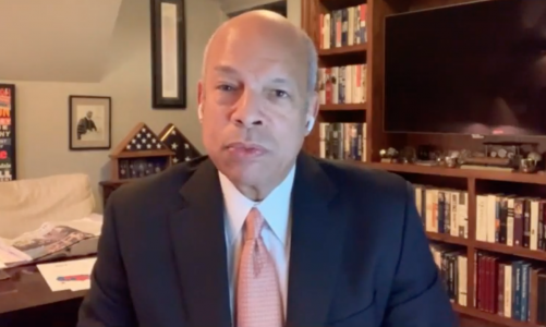 Bald, older, African American man (Jeh Johnson) in suit and tie sits in living room with airpods and serious expression.