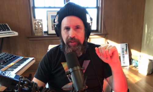 Bearded caucasian man (Duncan Trussell) looks into camera wearing hoodie and speaking into microphone.